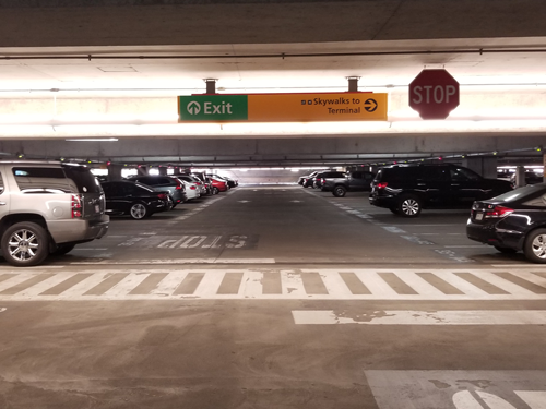 Square photograph taken inside the Hourly Garage at BWI Marshall Airport. Photo includes overhead wayfinding signage, a crosswalk and vehicles in parking spots.