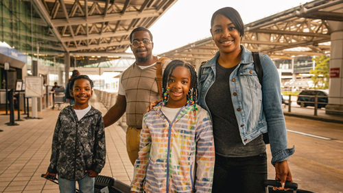 Photo of a family of four posing smiling as they hold carry-on bags along a terminal roadway at BWI Marshall Airport.