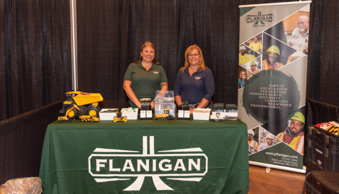 Two women posing for a photo at the Flanigan booth in the exhibit hall at the 2024 SynergyBWI Business and Networking Event.