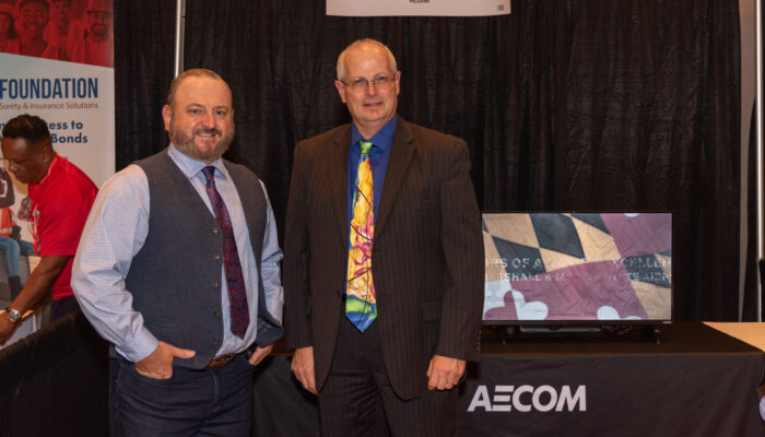 Two men posing for a photo in front of the AECOM booth in the exhibit hall at the 2024 SynergyBWI Business and Networking Event.