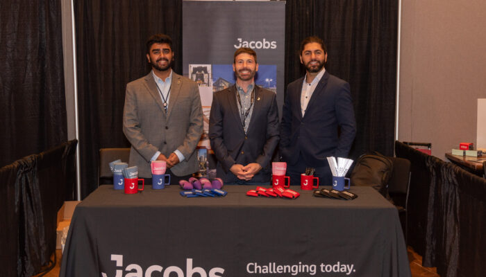 Three men posing for a photo at the Jacobs booth in the exhibit hall at the 2024 SynergyBWI Business and Networking Event.