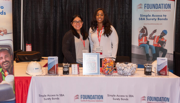 Two women posing for a photo at the Foundation Surety & Insurance Solutions booth in the exhibit hall at the 2024 SynergyBWI Business and Networking Event.