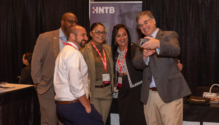 Five attendees posing for a photo in front of the HNTB booth in the exhibit hall at the 2024 SynergyBWI Business and Networking Event.