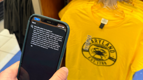 An individual holds a phone utilizing an assistance app with text describing a t-shirt on a rack inside a store at BWI Thurgood Marshall Airport.